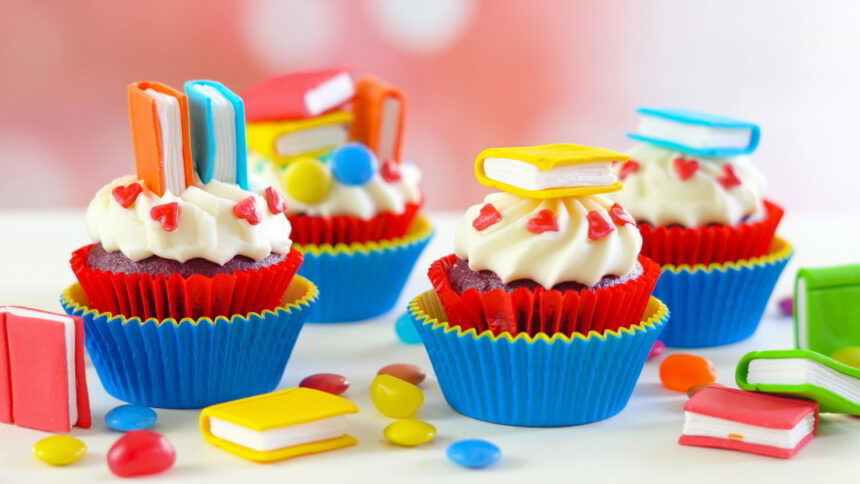 Book birthday chocolate cupcakes in layered red and blue papers sit on a table. Little fondant books sit atop stylishly piped white frosting.